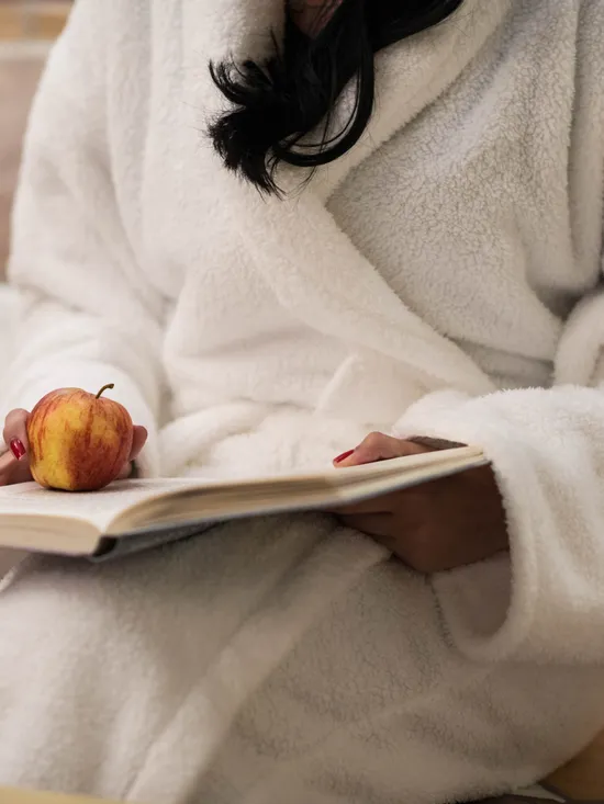 Frau im weißen Bademantel hält einen Apfel und liest ein Buch im Spa-Ruhebereich
